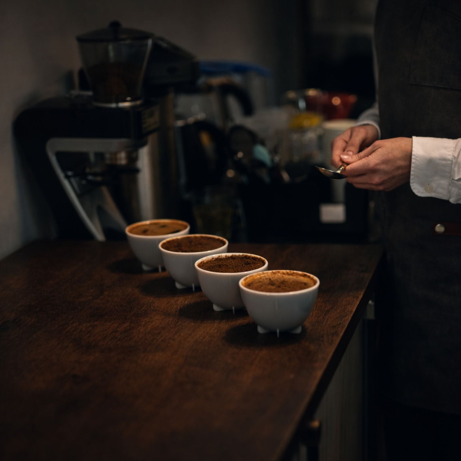 Coffee cupping setup with four white ceramic bowls on a dark wooden table, freshly brewed coffee grounds visible, as a taster prepares to evaluate aroma and flavor.